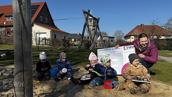 Kinder sitzen mit Erzieherin im Sand und halten Geschenke in der Hand